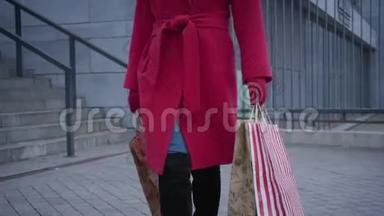 无法辨认的优雅女士穿着红色外套，手套和黑色靴子，带着购物袋在城市里散步。 时髦的年轻女子